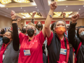 Four women in red scrubs, of various races, stand, all masked, raising their fists in the air, some clasping one another's hands. They are at the front of a large crowd in the ballroom.