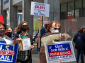 Picketers (Black and white, men and women) marching in front of a post office. Signs say "U.S. Mail Is Not for Sale," "Postmaster DeJoy Is Destroying Our Post Office," "Save Our Public Postal Service: Forever Essential." Also visible are copies of the flyer "Dump DeJoy and His 10-Year Plan."