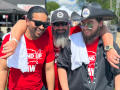 Three guys in red Stand-Up Strike (UAW) t-shirts, two bearded, stand outside on grass with arms around each other’s shoulders, smiling big.