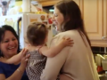 A white woman, right, hands a baby to a Latina woman, left. They are in an apartment kitchen. Both are smiling big. The baby's face is not visible.