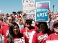 Workers in red UNITE HERE shirts pose at a huge rally. One handlettered sign says "Fair contract for Trump workers" and another printed sign says "Love Trumps Hate."