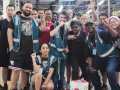 A group of workers, diverse in race and gender, poses together on the factory floor. Most appear young; one wears a prosthetic arm. A few have fists raised and most smile or wear determined expressions. Most wear shorts and some wear blue work vests. 