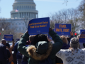 Crowd of people viewed from behind, holding blue AFGE sign. One says "Proud Government Worker, Proud Union Member. Another says "Stand, Unite, Fight." In front of them is the U.S. Capitol building.