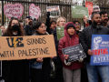 A chanting crowd stands in front of the White House fence. Three people are helping hold a large cardboard sign that says "UE stands with Palestine" with the union's lightning-bolt logo and a slice of watermelon. Other printed signs say "American Postal Workers Union: Fighting for Justice" and "Biden, you are starving Gaza. Permanent ceasefire now!" 