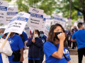 People in blue "INA" shirts march. Some carry picket signs "Safety in Numbers," "Respect Nurses!" "Unfair Labor Practice Strike." One speaks into a bullhorn mic.