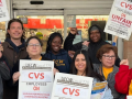 Eight people, varied in race and gender, pose together in front of a CVS store. Some wear UFCW yellow shirts and most carry picket signs that say "UFCW Local 770: CVS employees on unfair labor practices strike, please respect the picket line" in English and Spanish.