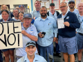 Seventeen uniformed letter carriers pose together in a post office, holding a big handmade "VOTE NO!" sign.