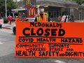 masked worker in front of mcdonald's holds big orange sign: "MCDONALD'S CLOSED, COVID HEALTH HAZARD, COMMUNITY SUPPORTS WORKERS STRIKE FOR HEALTH, SAFETY AND DIGNITY"