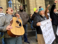 A group of people stands outside a building, singing. Edwin, a young man with guitar, has his head leaned back shouting into the sky. Several other people have fists in the air, one wears a big smile, and some hold big paper on which lyrics are written out: "Sign me up for the union, bring me my union card, We can work better with decent pay, health care, childcare, and more of a say..."