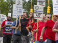 A group of people in red shirts, one man carrying a child march past the camera with signs saying Stellantis is barganing in bad faith.