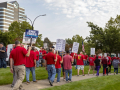 Workers in red shirts picket outside. Many of the picket signs are white with UAW logo, Local 412, and the words "Stellantis Bargaining in Bad Faith."